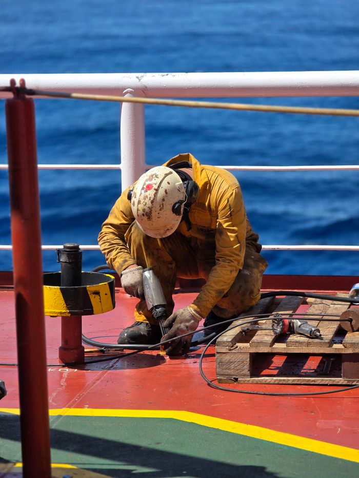 Worker in protective gear using tools on a ship deck against ocean backdrop. Labor and safety at sea.